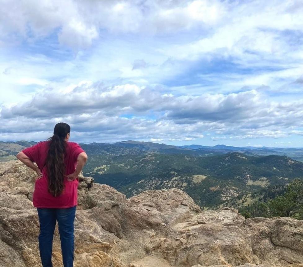 Ms. Only A Chief faces away from the camera, overlooking green, wooded mountains under a blue sky.
