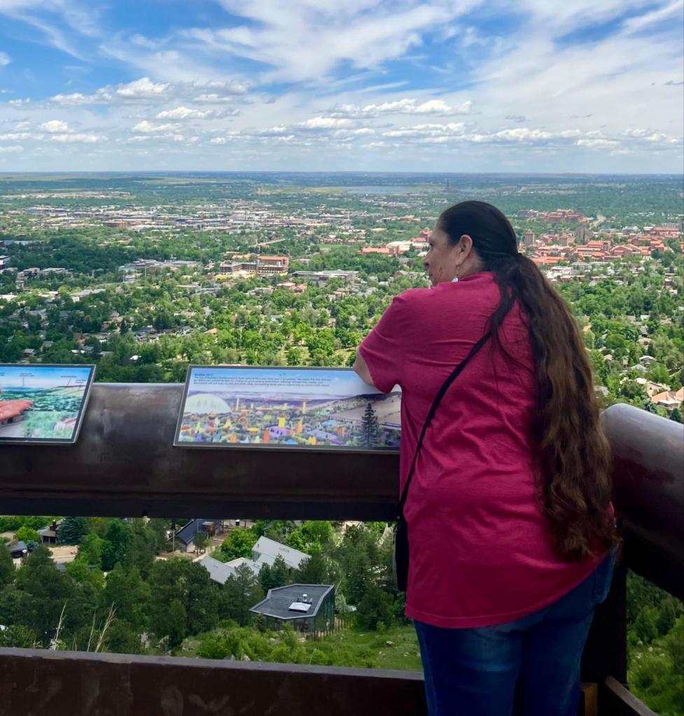 Ms. Only A Chief looks out over the University of Colorado Boulder campus from an OSMP overlook, with blue sky, white clouds, and red‑roofed buildings in the distance.