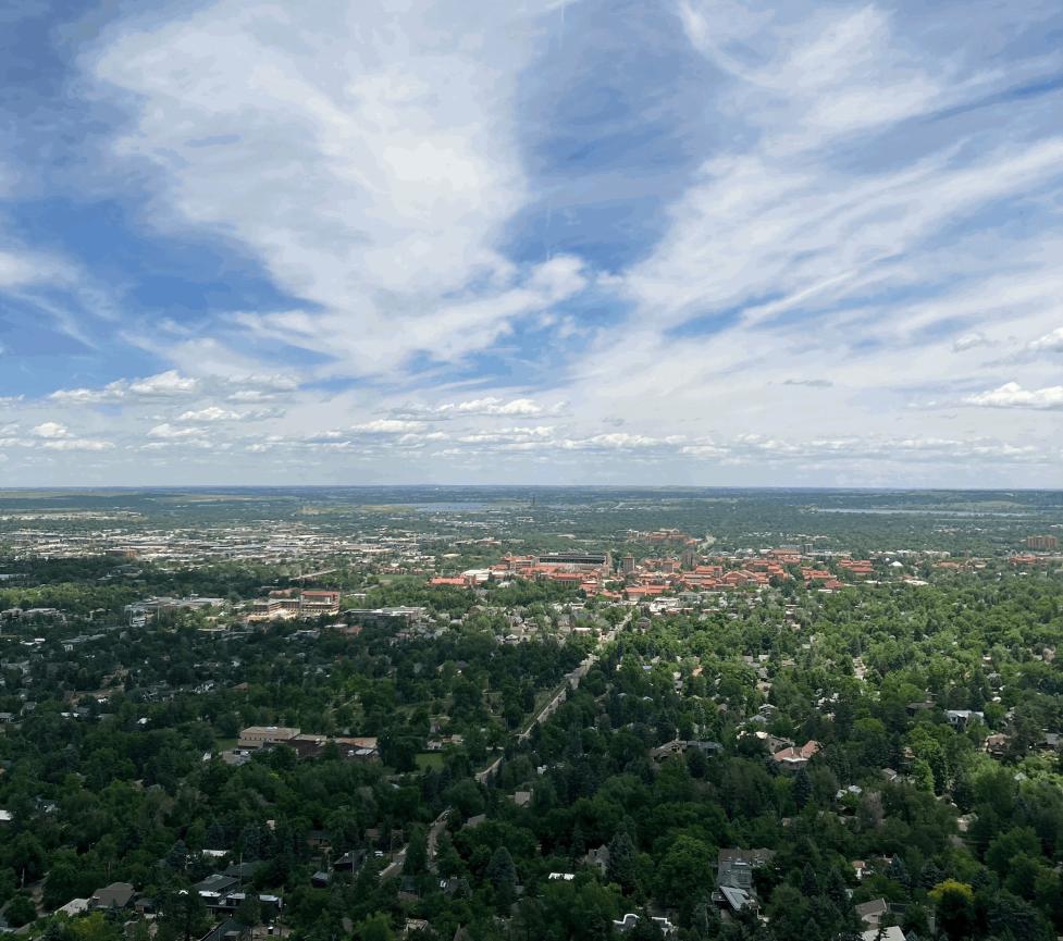 Boulder OSMP lookout with CU campus, red roofs amid green trees, blue sky with wispy clouds.