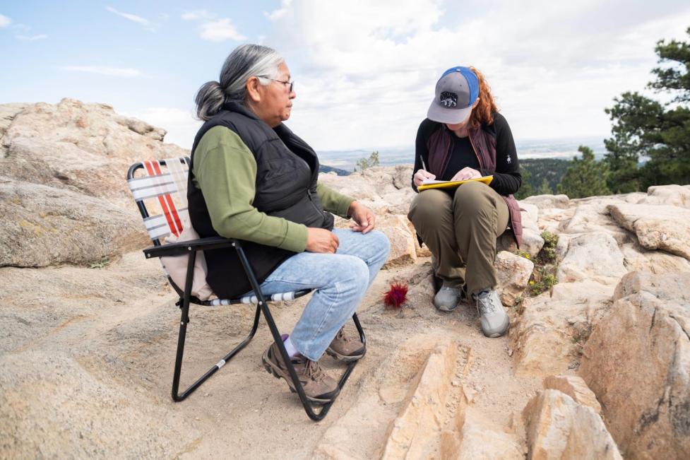 Ms. Cindy Badback and Jessica Yaquinto sit in a rocky area; Ms. Badback speaks while Jessica looks down at her notes.