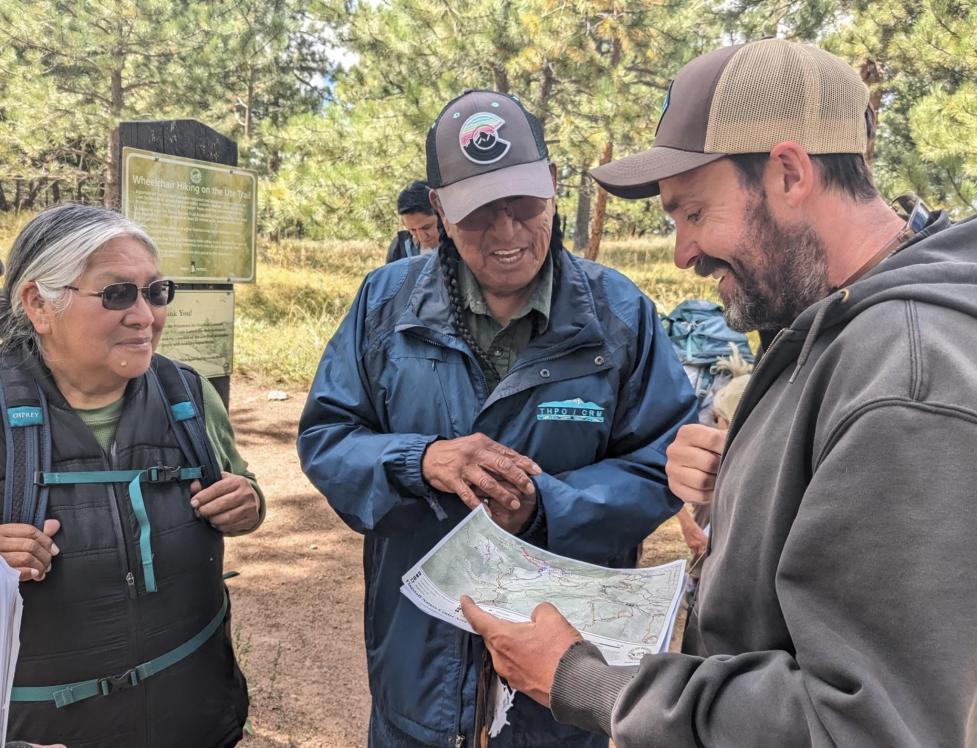 Three people stand in a semicircle near a trailhead sign, laughing as they look over a map.
