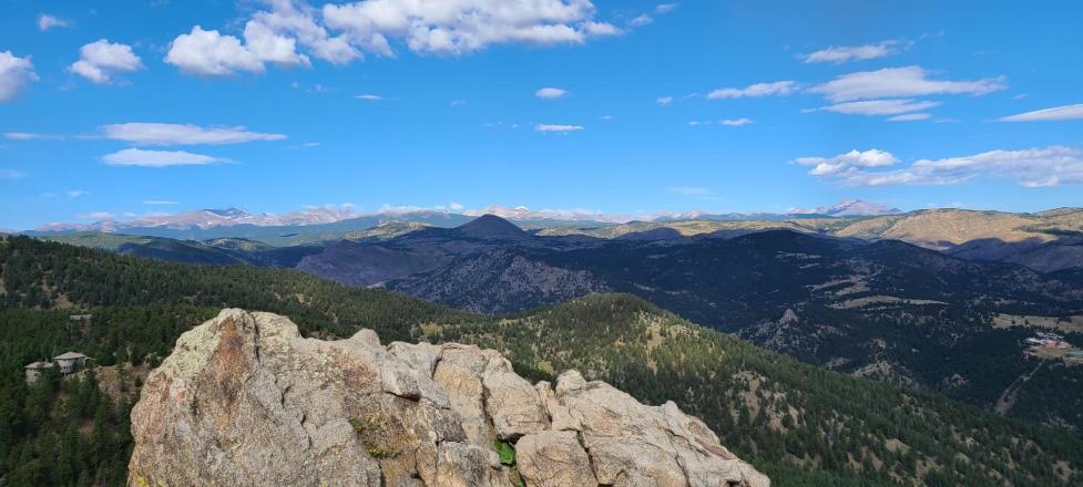 View from a Boulder OSMP trail with large rocks in the foreground, green conifer‑covered mountains in the midground, and snow‑topped peaks in the distance.