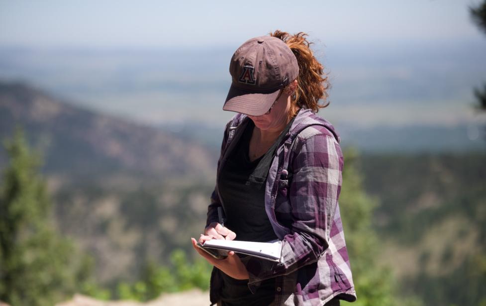 Ethnographer Jessica Yaquinto stands in the foreground, writing in a notebook, with a blurred mountainous landscape behind her.