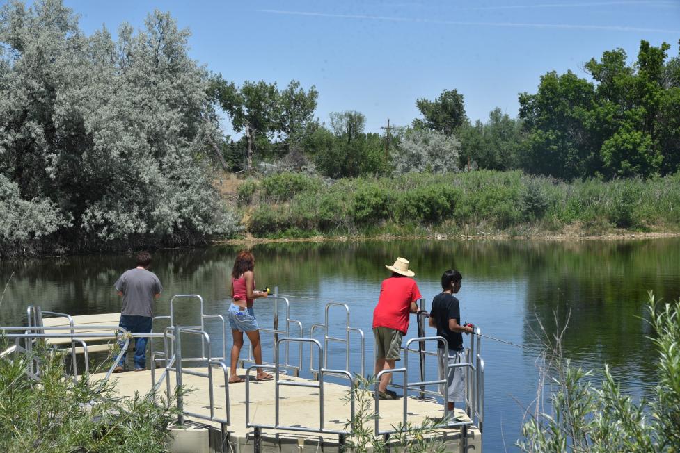 Three people standing on a pier holding fishing poles while looking out a pond surrounded by greenery.