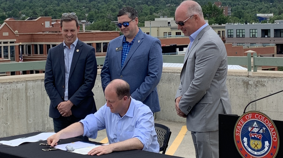 Governor Polis, seated, signs a bill on a Boulder rooftop, surrounded by Colorado state legislators. 