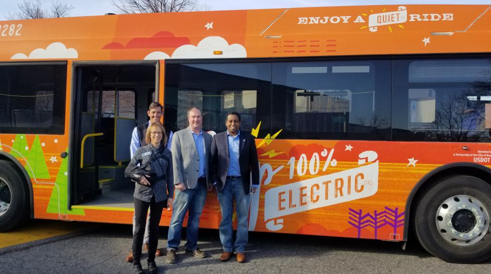 Government officials stand in front of an orange, all-electric city bus