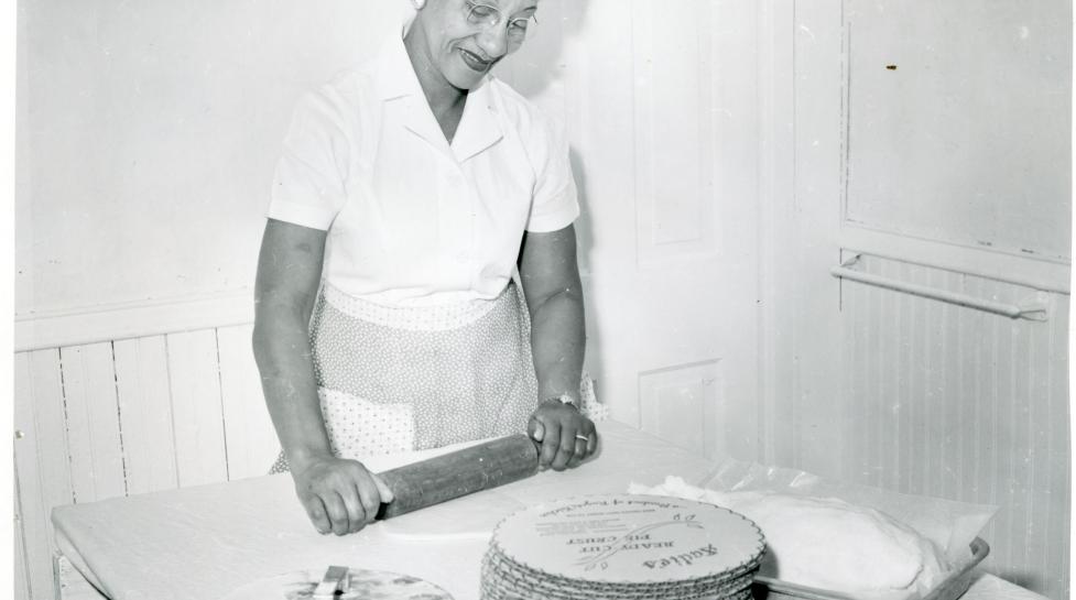 Sadie Arnold, a woman with short black hair and white-rimmed glasses, rolling out dough besides a stack of flat pie crusts.