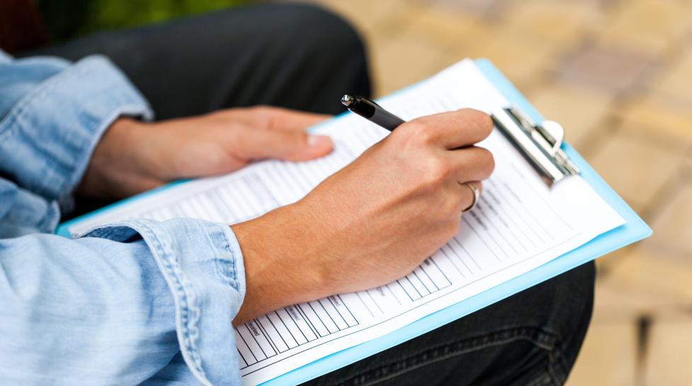 Person sitting down writing on a clipboard that is resting on their knee