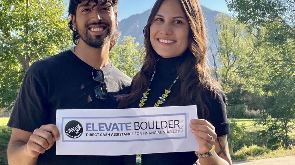 Two people smiling at the camera holding a sign with the Elevate Boulder logo.