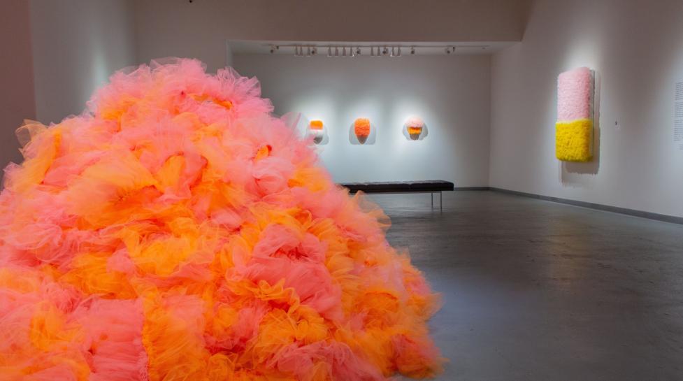 Orange and pink tulle fabric sewn together into an abstract sculpture on the floor of an art gallery.