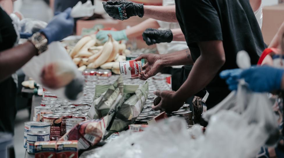 Scene from a food bank, picture shows the hands of volunteers over a table full of various food items. 