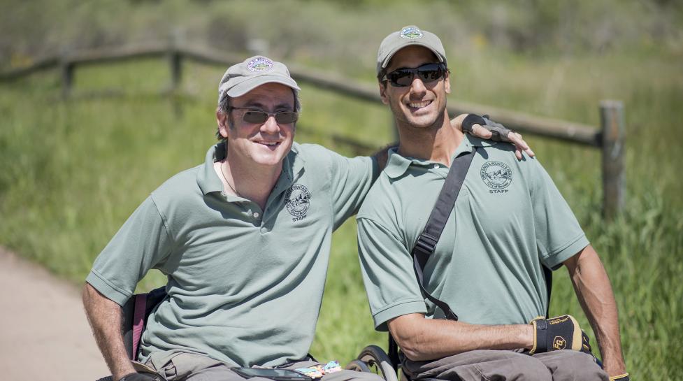 Two wheelchair users who are City of Boulder employees smiling at the camera while enjoying a roll on an accessible trail. 