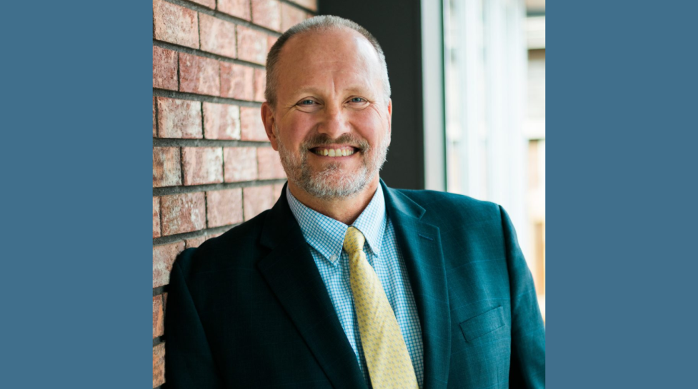 A headshot of Dave Ruppel. In the photo, Ruppel is a middle-aged white man with graying hair and a beard. He is smiling at the camera and wearing a suit with a yellow tie and blue dress shirt.