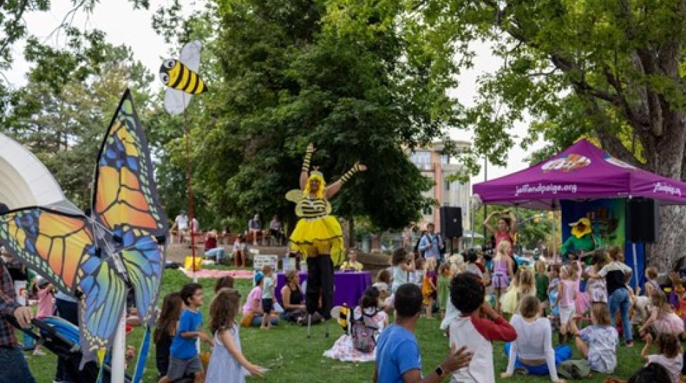 Outdoor festival scene at a park with children and families watching a performer.