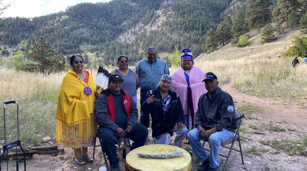 Tribal elders seated around a large drum with the Boulder mountains in the background.