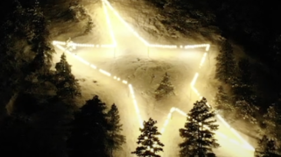 Large star made from lights on the side of a snowy mountain with pine tree around it.