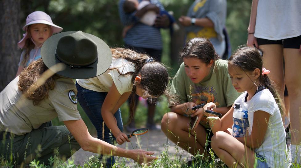 Group of young kids knealing around a Ranger in a khaki outfit with a hat looking at piece of grass.