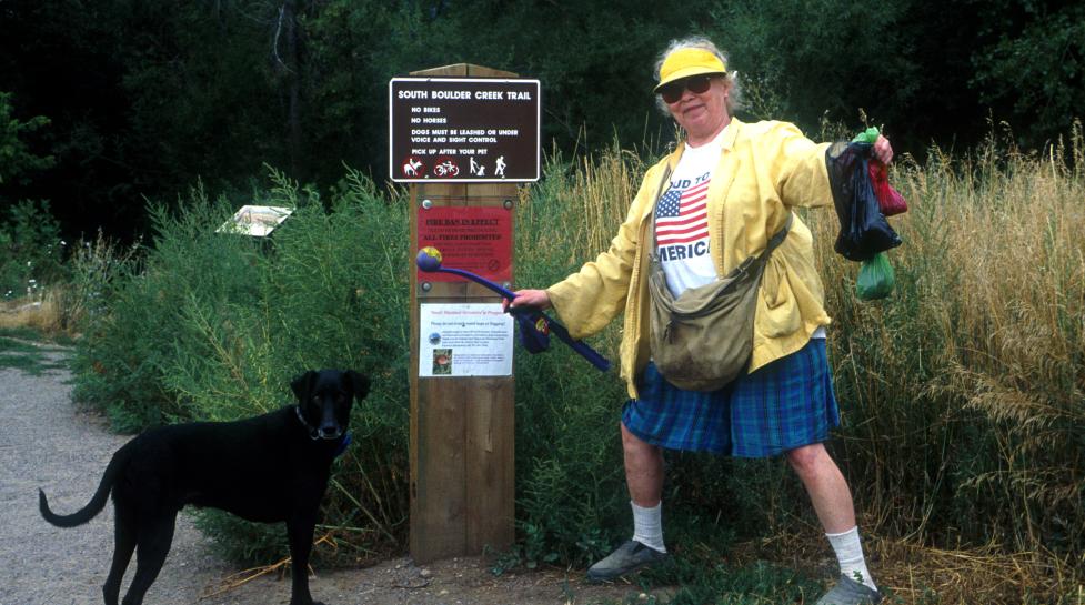 Woman carrying two poop bags to a trash can while holding her dog on leash.