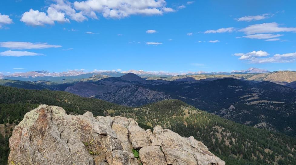 View from a Boulder OSMP trail with large rocks in the foreground, green conifer‑covered mountains in the midground, and snow‑topped peaks in the distance.