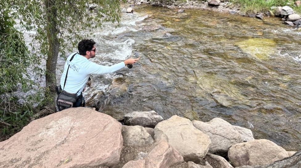 Man with arm stretched out to cast a fishing line over a full river with trees and rocks surrounding the river.