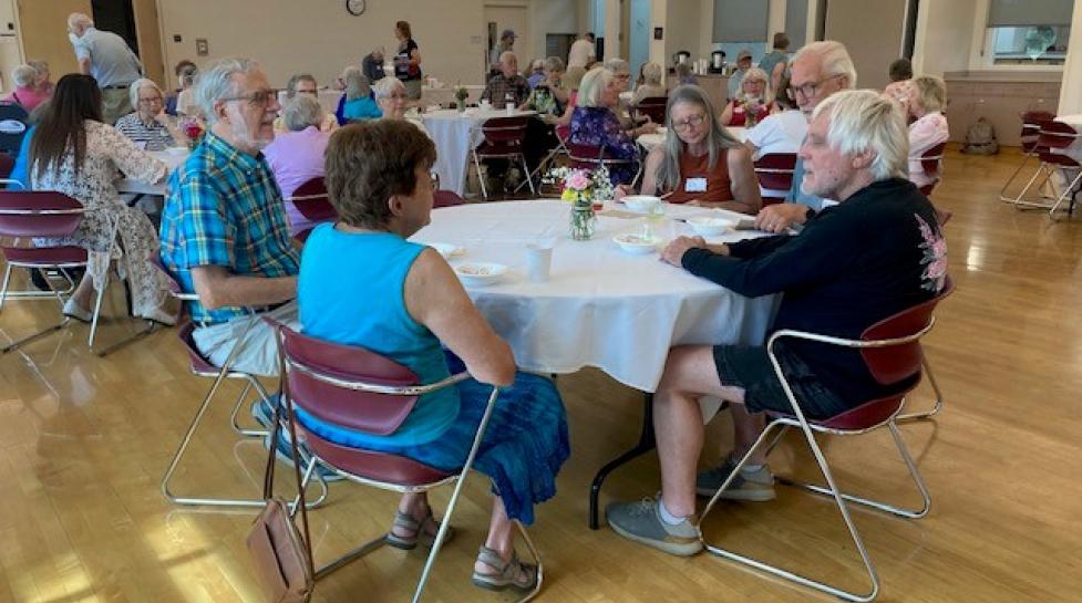 Groups of older adults sitting around tables conversing, in the East Age Well Center. 