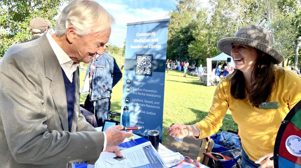 A man reviews materials at the Community Mediation and Resolution Center information table while a smiling woman in a yellow shirt assists him at a sunny outdoor information fair.