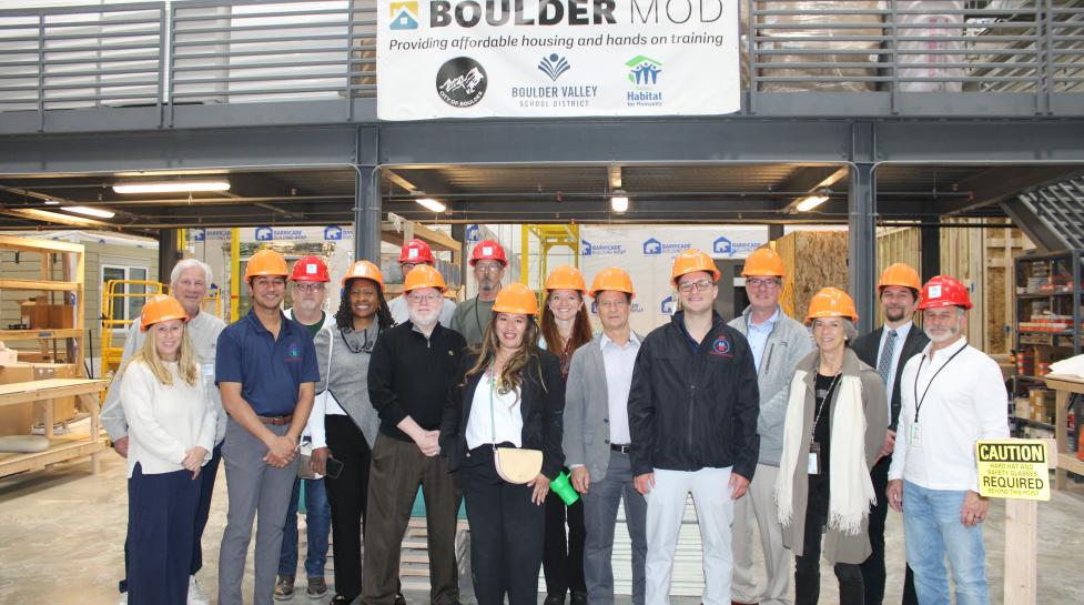 A group of approximately 15 adults wearing orange hard hats pose for a group photo inside BoulderMOD. 