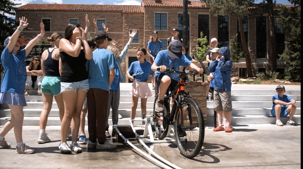 kids in blue shirts with hands outstretched to catch water as one kid pedals on a bike