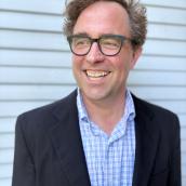 Headshot of Blythe Bailey in black suit jacket, blue plaid shirt and glasses