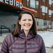 White woman with shoulder length brown hair who is smiling and wearing a purple jacket while standing in front of building.