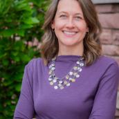 Woman Smiling in Purple long sleeved shirt in front a an exterior brick wall and tree.