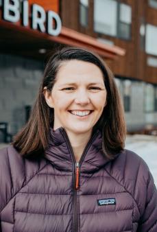 White woman with shoulder length brown hair who is smiling and wearing a purple jacket while standing in front of building.