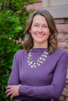 Woman Smiling in Purple long sleeved shirt in front a an exterior brick wall and tree.