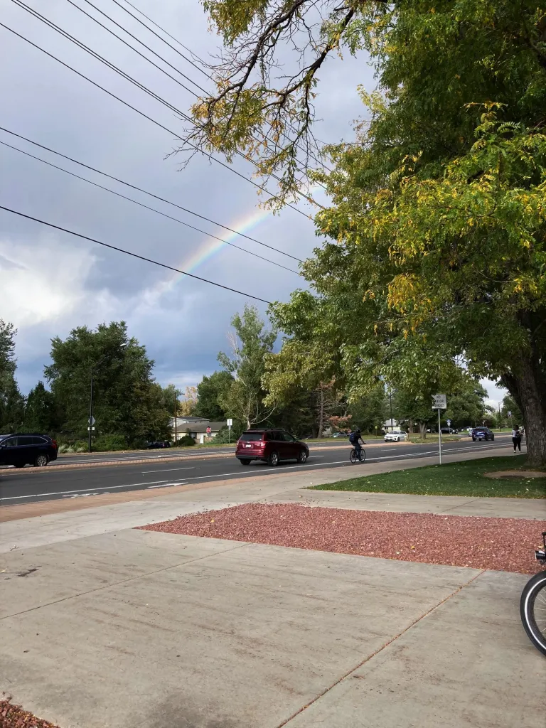 A rainbow near a road. There is a sidewalk and a large driveway that leads to it.