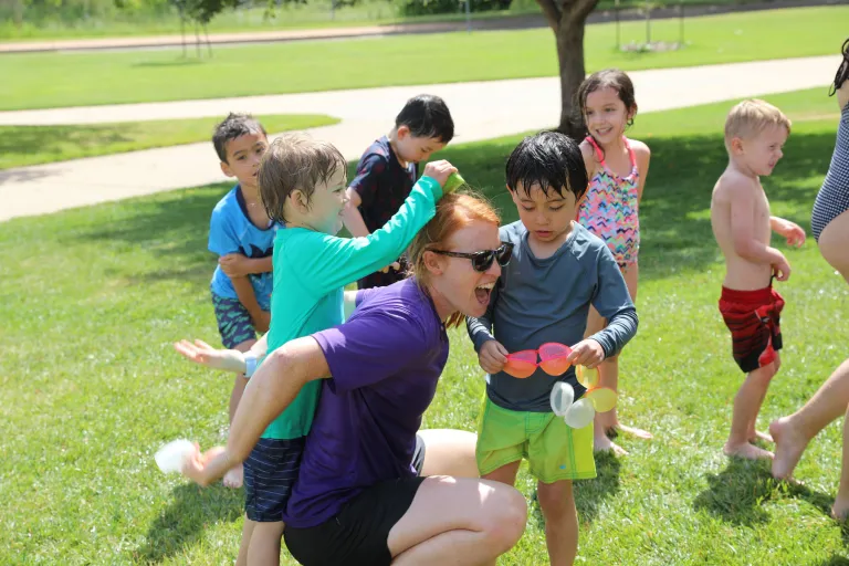 Smiling children pile onto a camp counselor outdoors at one of Boulder’s Kidz Kamp opportunities.