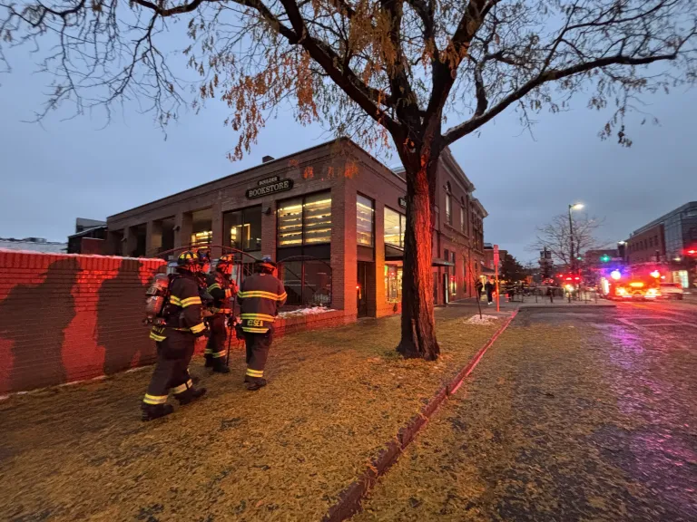 Four Boulder firefighters dressed in full gear approaching the Boulder Bookstore at dusk lit up by the blue and red lights from nearby vehicles. 