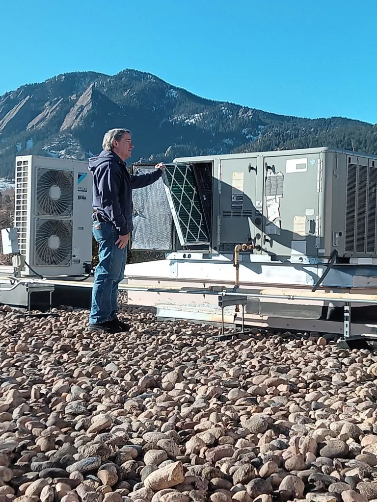 A facilities technician repairing a heating/cooling unit on the rooftop of the Penfield Tate II Municipal Building with the Flatirons in the backdrop.