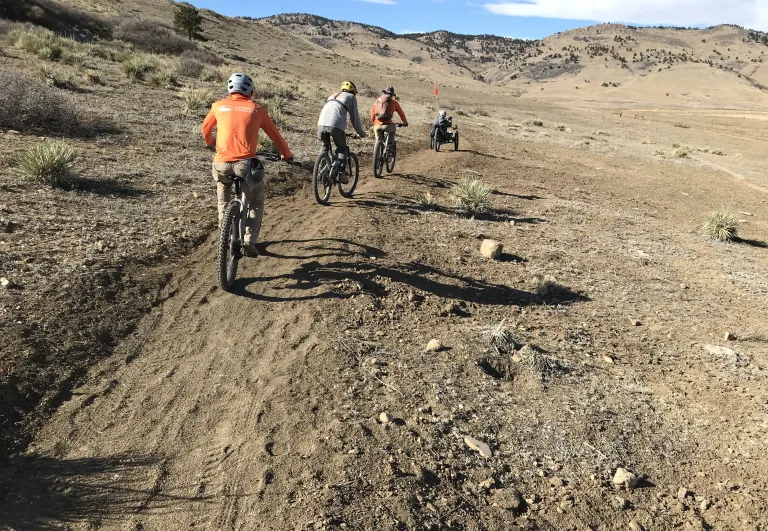 A group of cyclists riding on the single-track Cobalt Trail through brown, winter grass under a sunny, blue sky.