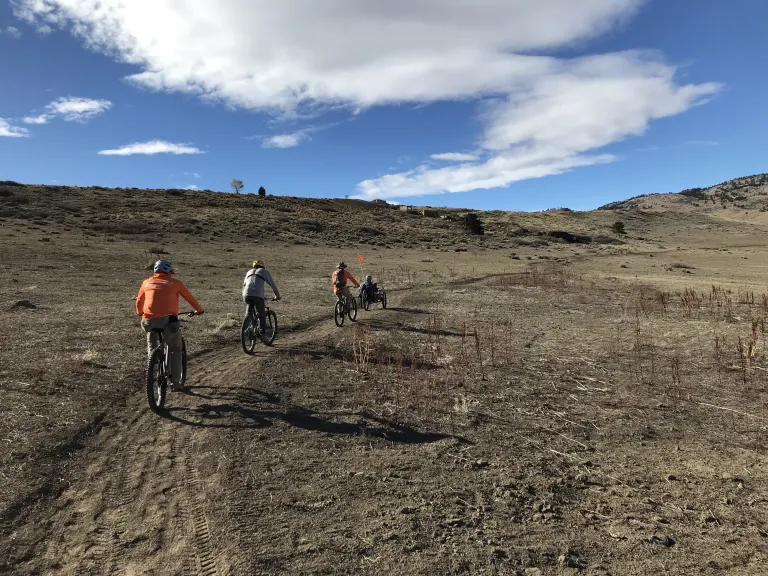 A group of cyclists riding on the single-track Cobalt Trail through brown, winter grass under a sunny, blue sky.