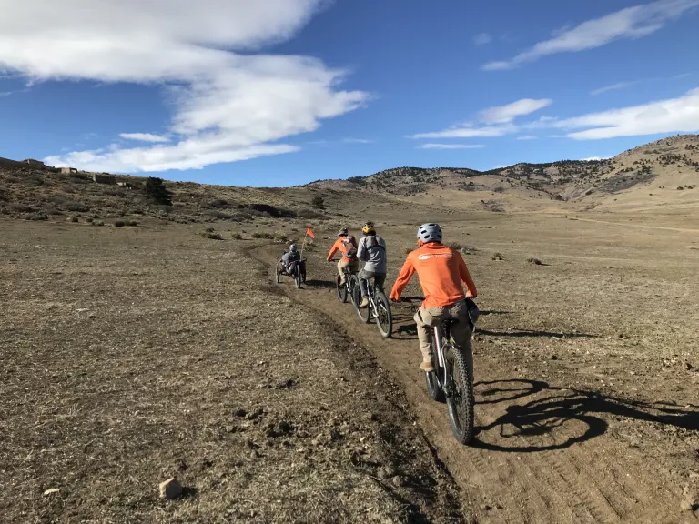 A group of cyclists riding on the single-track Cobalt Trail through brown, winter grass under a sunny, blue sky.