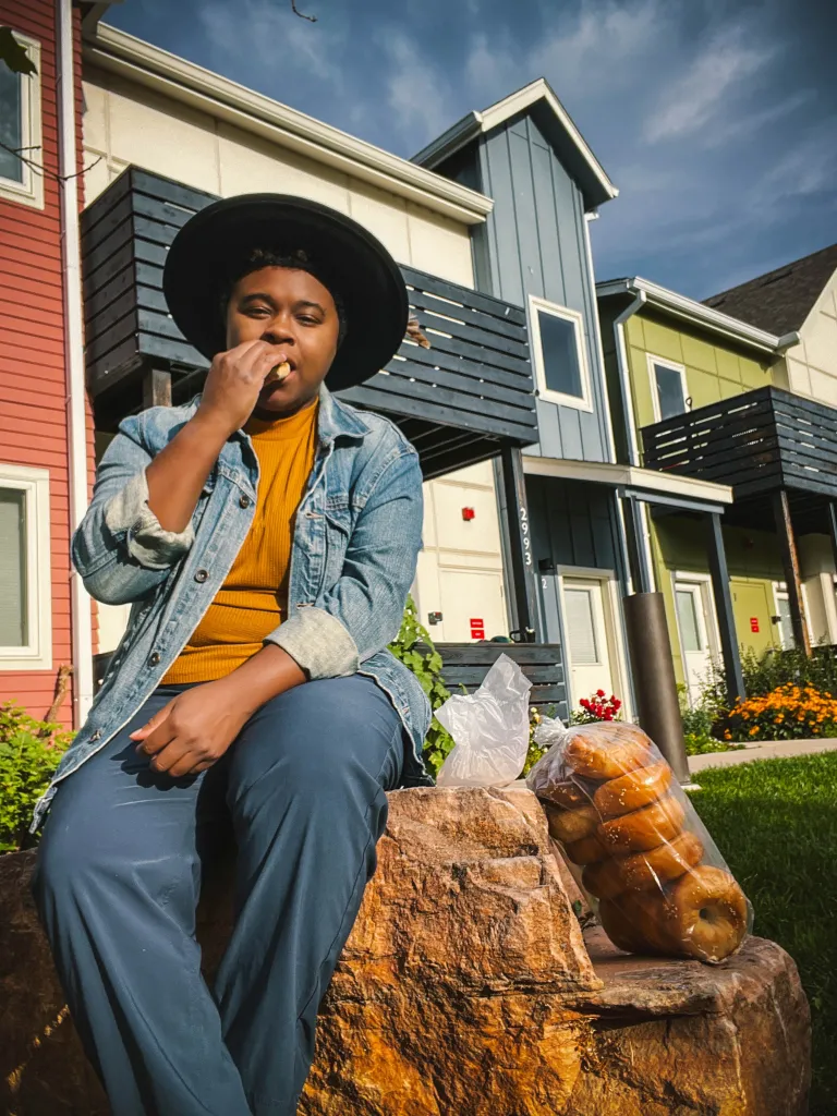 Participant sits on a rock eating a bagel. A bag of bagels sits next to the participant. There are homes in the background. 