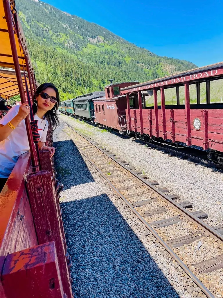 Woman leans head out of a train car. There is another train in the background and there are mountains in the background behind the trains. 
