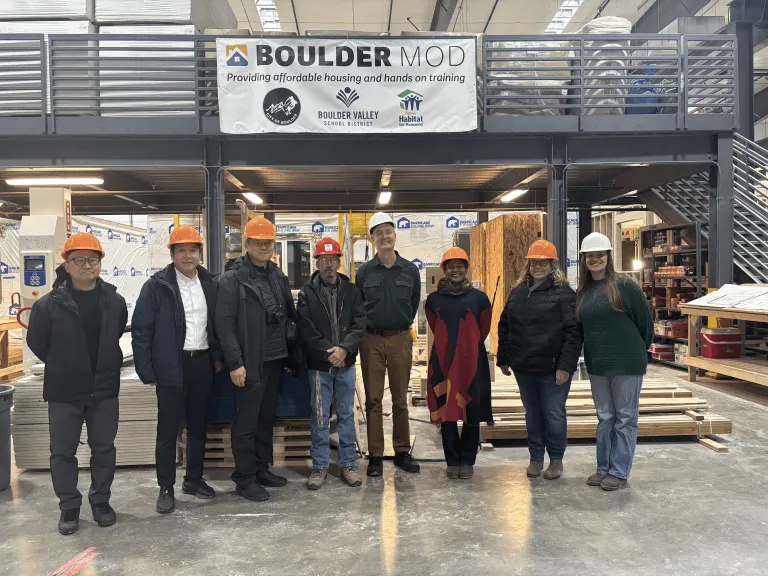 A delegation from the Japan Off-site Construction Association, Councilmember Taishya Adams and Habitat and city staff pose in front of the BoulderMOD sign on the factory floor.