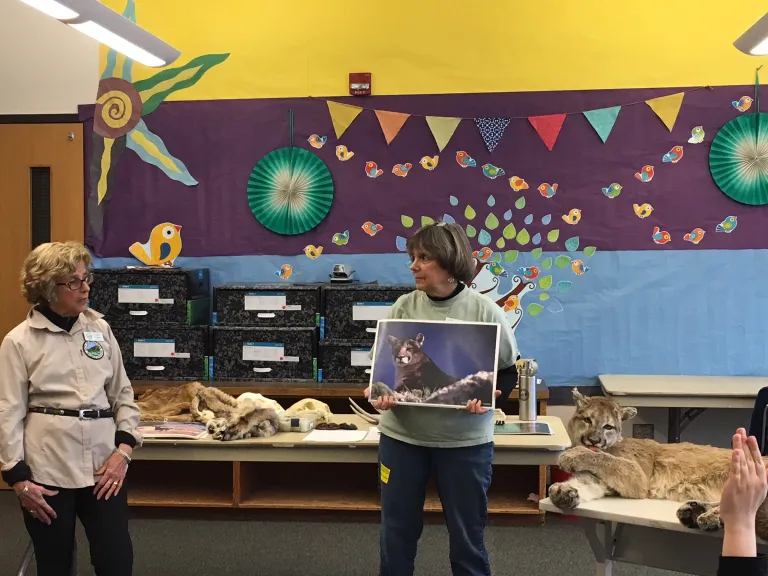 Two adult women standing in a colorful classroom pointing at animal photos, skulls and pelts