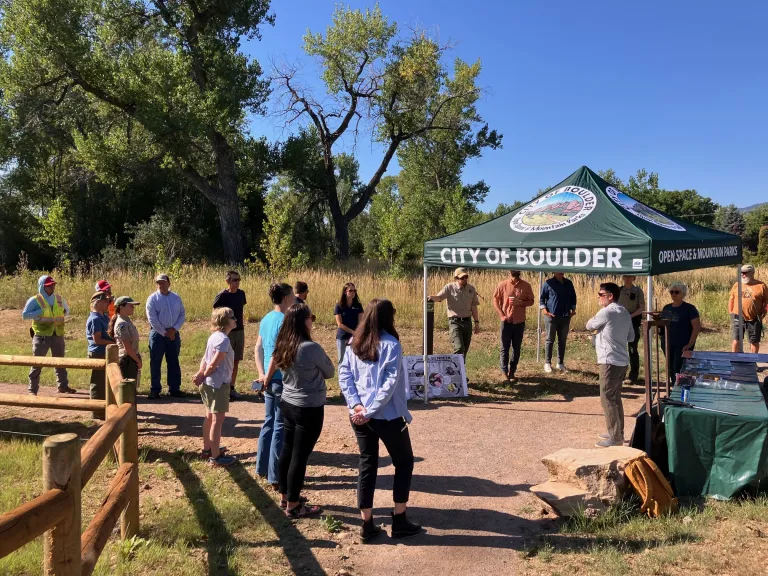 People standing around a city of boulder tent on a trail outside.
