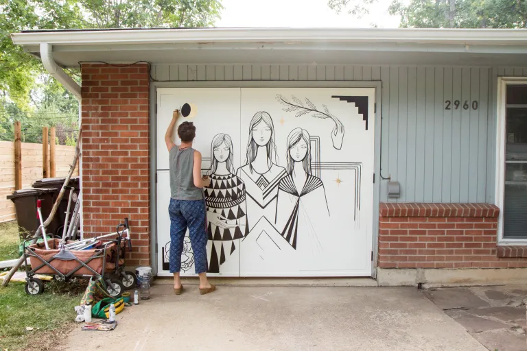 Artist painting a mural of women on the garage door of a house