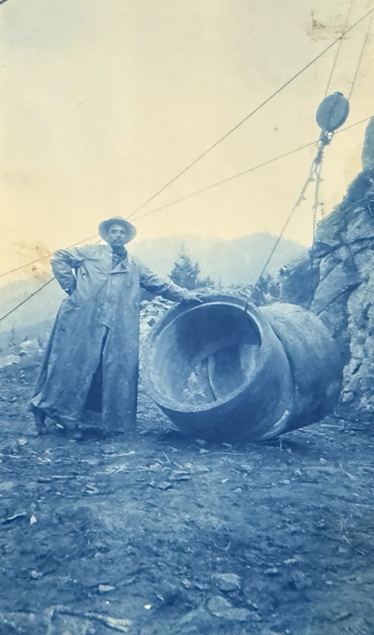 Undated, a man standing near sections of pipe being hoisted by the tram system. 