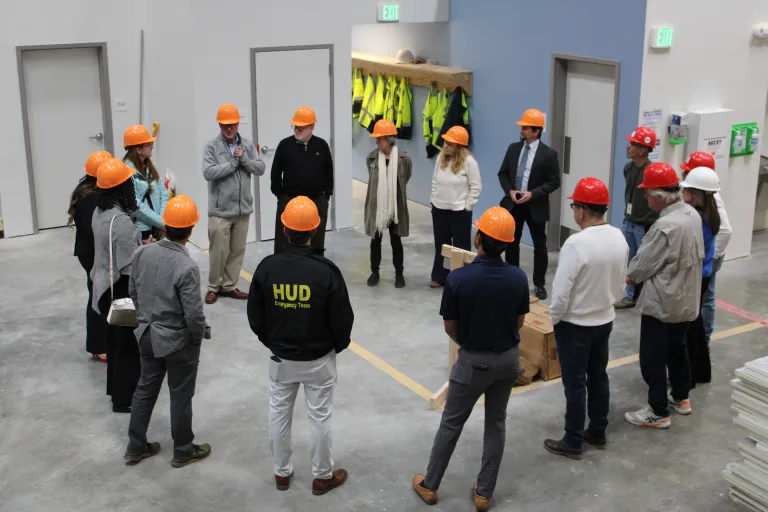Approximately 15 tour participants wearing orange and red hard hats gather in a loose circle inside BoulderMOD for a group discussion. 