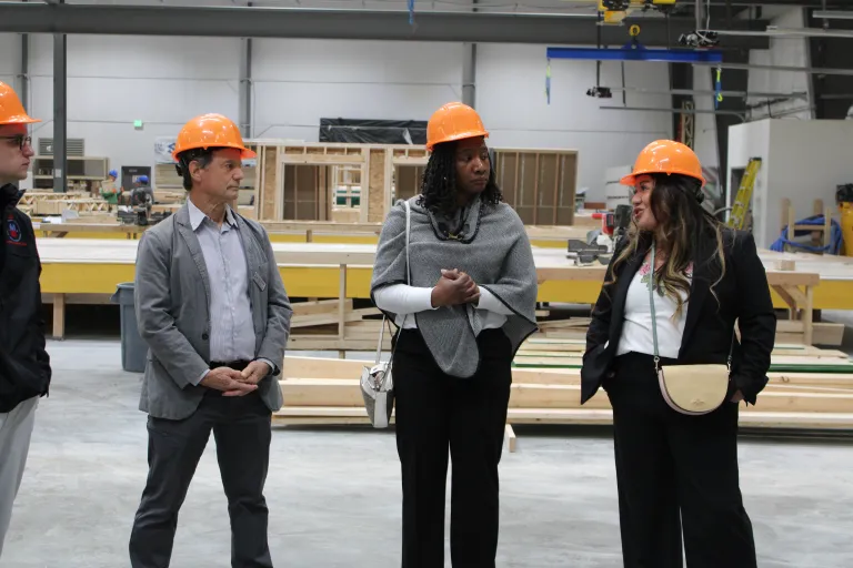 Three visitors wearing orange hard hats converse on the floor of the BoulderMOD, with partially framed modular housing panels and construction equipment visible in the background.