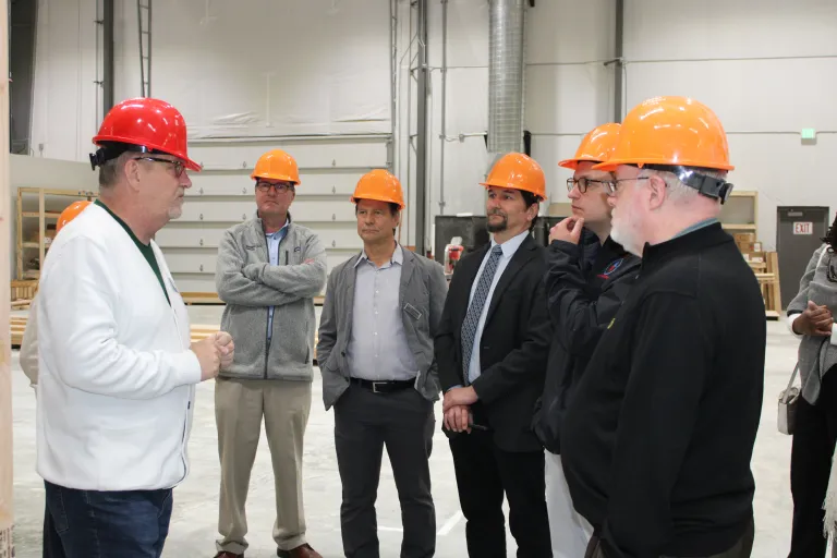 A person in a red hard hat and white jacket leads a discussion with five visitors wearing orange hard hats inside BoulderMOD.